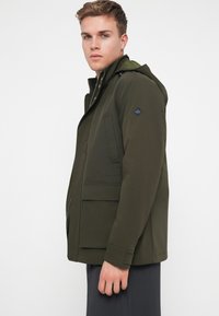 Olivovo zelená bunda s kapucňou, s uzatváraním na zips, plochými vreckami a štruktúrovanou tkaninou. Obsahuje nášivku s logom na rukáve.