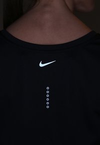 Musta Nike-urheilupaita, jossa on heijastava logo kauluksessa ja kuusi tasaisesti sijoitettua heijastavaa ympyrää selän keskiosassa.
