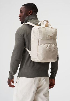 Beige Rucksack mit gepolstertem Design, Obergriffen und verstellbaren Trägern. Glatte Textur mit zwei runden Akzenten auf der Vorderseite.