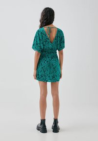 Robe à manches courtes de couleur turquoise avec un motif floral noir, taille cintrée et dos dégagé. Associée à des bottines noires à la cheville. Texture de tissu lisse.