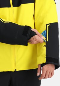 Wasserabweisende Jacke mit einem leuchtend gelben und schwarzen Farbschema, Reißverschluss vorne, Klettverschlussmanschetten und einer Karten Tasche am Ärmel.