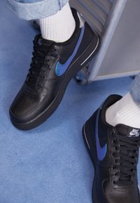 Person som bär svarta läder Nike Air Force 1 sneakers med iriserande blå swoosh och vita strumpor, sittande på ett blått heltäckningsmatta.
