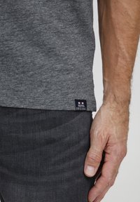 Šedé tričko s krátkym rukávom, s jemnou textúrou a miernym melírovaním; obsahuje malé označenie značky na leme, kombinované s tmavými džínsami.