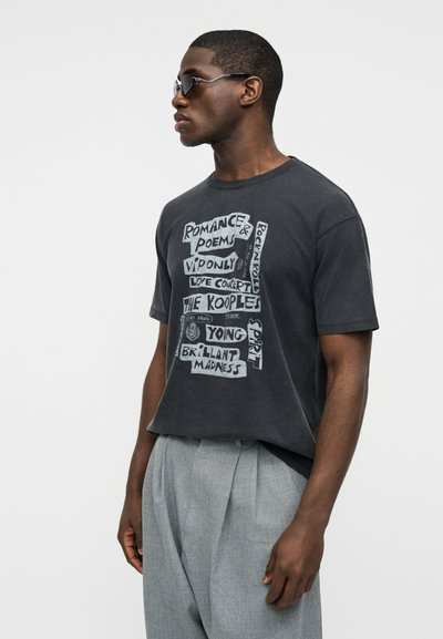 Άνδρας που φοράει μαύρο T-shirt με γράμματα, γκρι ψηλόμεσο παντελόνι και μαύρα γυαλιά ηλίου, κοιτάζει προς τα αριστερά μπροστά σε ένα απλό ανοιχτό φόντο.