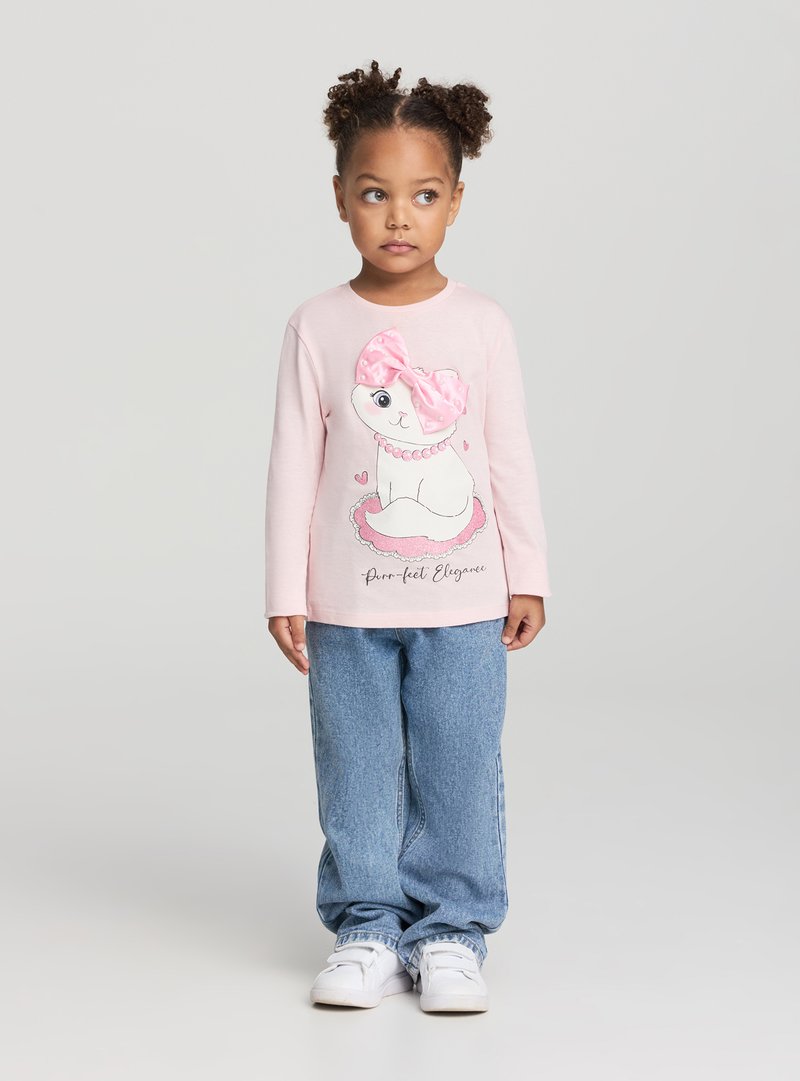 Maglietta a maniche lunghe rosa chiaro con una stampa di un gatto con un grande fiocco rosa. Abbinata a jeans blu e sneakers bianche, in piedi davanti a uno sfondo grigio.