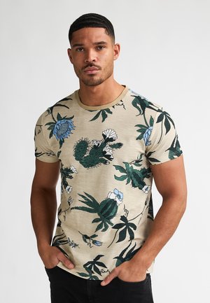T-shirt beige à motifs floraux en coton, présentant des motifs de plantes verts et bleus, col rond, manches courtes, et une finition texturée.
