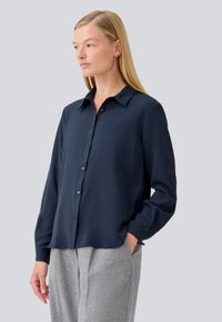 Camicia a maniche lunghe blu navy con colletto a camicia, caratterizzata da una texture liscia e una vestibilità comoda, abbinata a pantaloni grigi a spina di pesce.