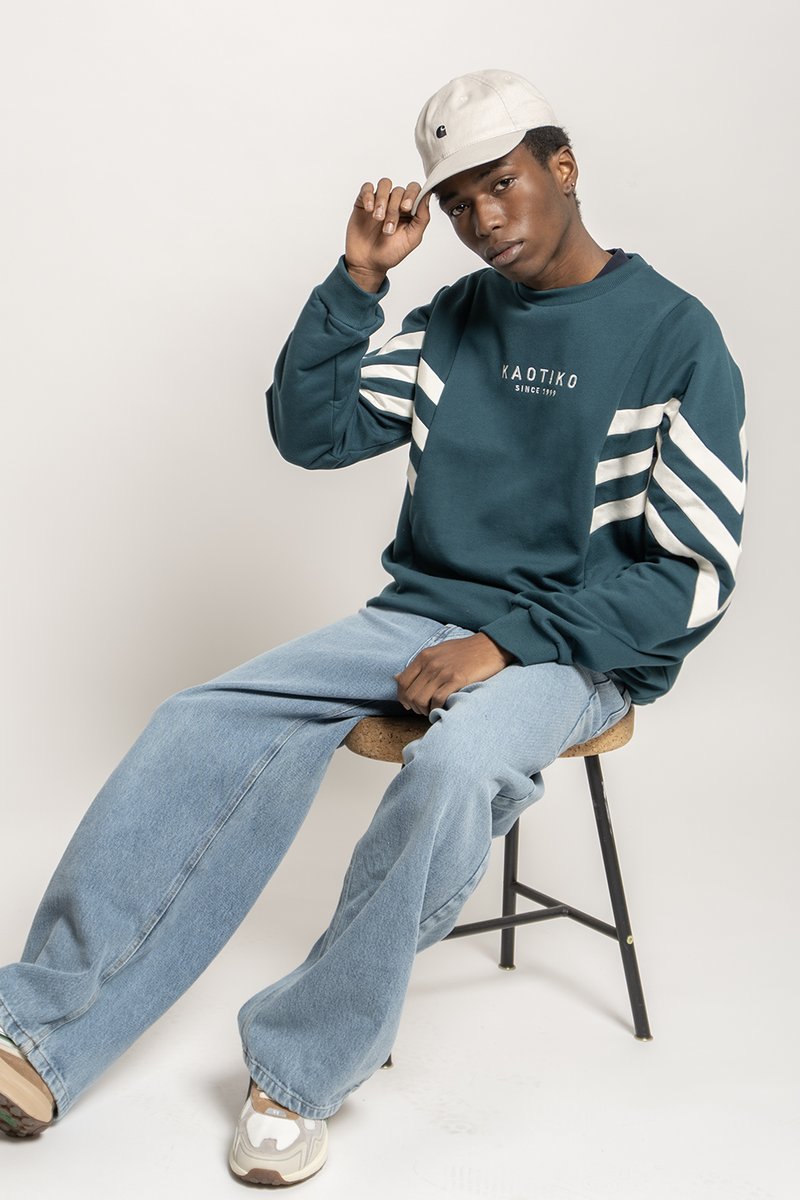 Teal sweatshirt med hvide striber, med "KAOTIKO" logo. Lyseblå brede jeans og en beige kasket. Uformelt fodtøj.