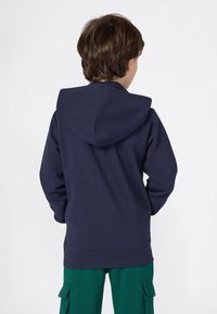 Bambino con i capelli corti castani che indossa una felpa blu navy e pantaloni verdi con tasche cargo, in piedi di spalle su uno sfondo bianco.