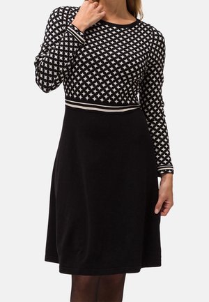 Femme portant une robe noire avec un haut à motif géométrique blanc et une jupe noire unie, touchant son cou d'une main.