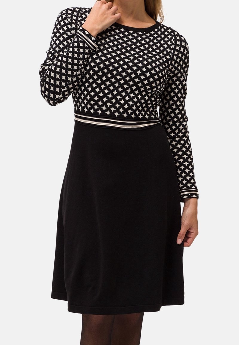 Femme portant une robe noire avec un haut à motif géométrique blanc et une jupe noire unie, touchant son cou d'une main.