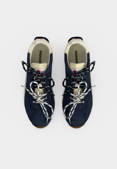 Paire de baskets bleu marine et blanc avec des lacets noirs et blancs mélangés noués en nœuds, vue de dessus sur fond blanc.