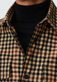 Chemise en flanelle à carreaux marron et noire avec un col roulé noir en dessous. Elle présente un col classique et des poches avant à boutons.