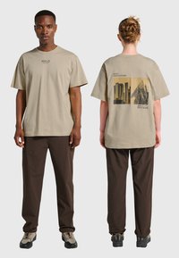 Beige bomullst-shirt med ett grafiskt tryck på baksidan, kombinerad med bruna byxor. Designen innehåller text och geometriska mönster.