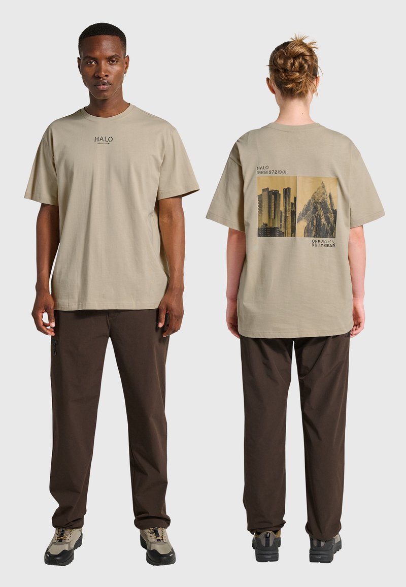 Beige bomullst-shirt med ett grafiskt tryck på baksidan, kombinerad med bruna byxor. Designen innehåller text och geometriska mönster.