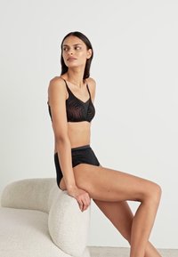 Ensemble de lingerie noire comprenant un soutien-gorge texturé à fines bretelles et un slip taille haute assorti, exposé sur une chaise de couleur claire.