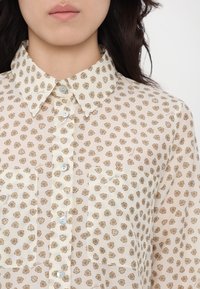 Sheer beige blouse with a small floral print, button front, collar, and two chest pockets. Subtle texture, lightweight fabric.