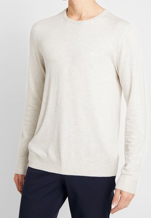 Mand iført en lysebeige langærmet sweater og mørke navy bukser mod en ensfarvet baggrund.