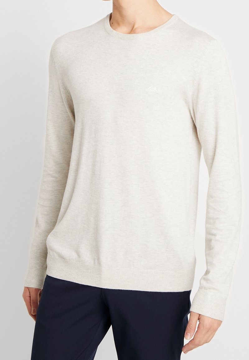 Homme portant un pull beige clair à manches longues et un pantalon bleu marine foncé sur un fond uni.