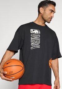 Μαύρο υπερμεγέθες t-shirt με λογότυπο NBA σε λευκό και γραφικό πορτοκαλί μπάσκετ, συνδυασμένο με πορτοκαλί μπάλα μπάσκετ, φορεμένο με κόκκινα σορτς.