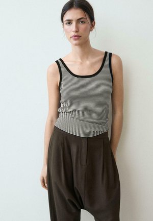 Femme portant un débardeur rayé noir et blanc et un pantalon noir ample, debout devant un fond clair uni.