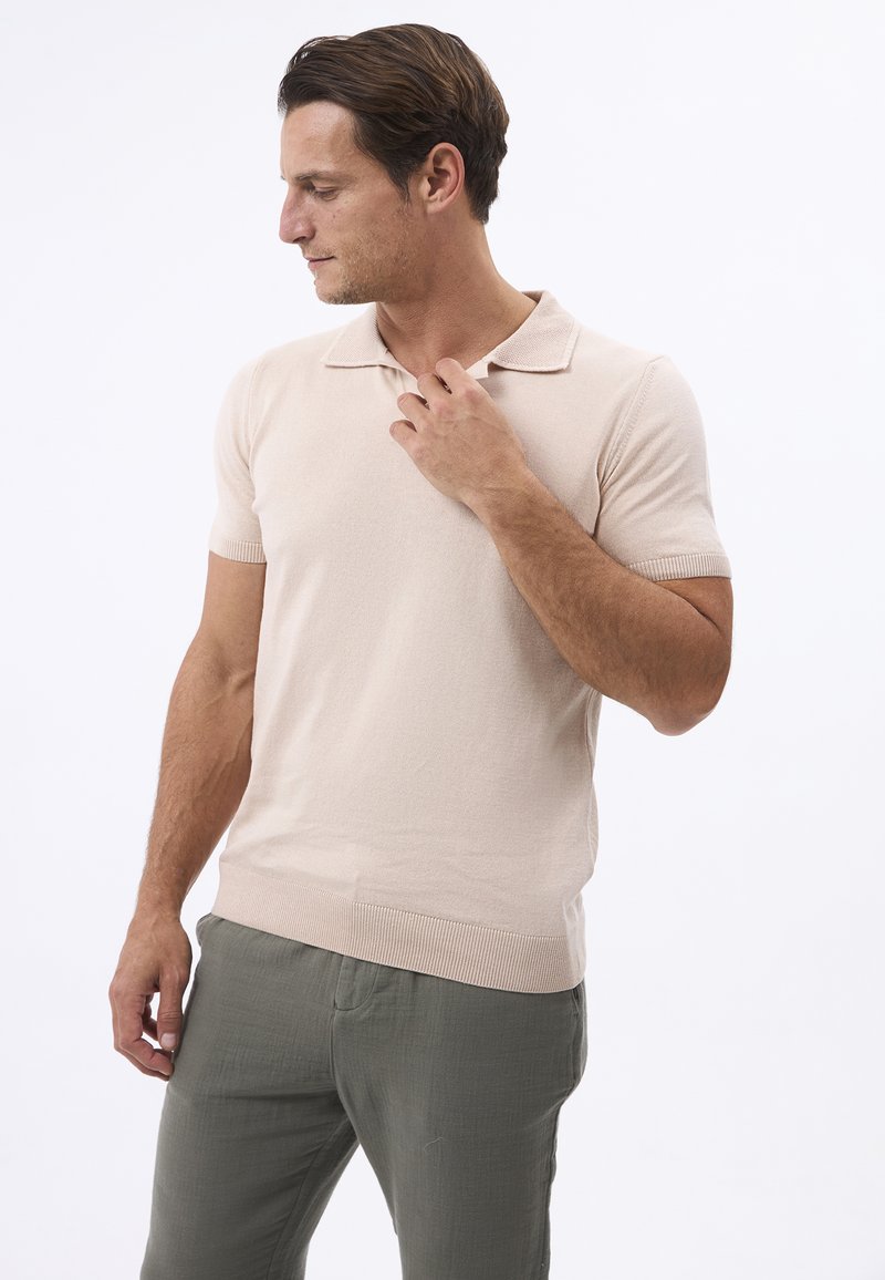 Polo shirt beige a maniche corte in un tessuto morbido con colletto, caratterizzato da orlo e polsini a costine. Indossato con pantaloni chiari e texture.