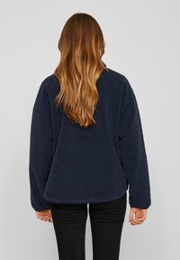 Moteris su ilgais rudiem plaukais, vilkinti tamsiai mėlyną flisinę striukę ir juodas kelnes, stovi nugarą į kamerą prieš paprastą foną.