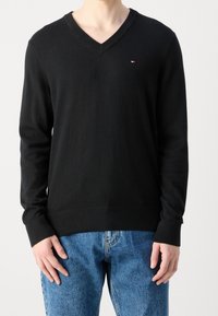 Musta V-kaula-aukkoinen neulepusero, jossa on pitkät hihat, teksturoitu neuloskangas, resorit hihansuissa ja helmassa, vasemmalla rinnalla pieni logo.