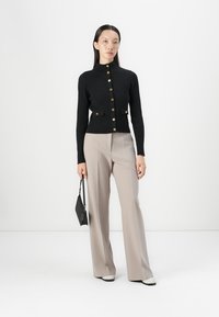 Cardigan nero a coste con bottoni dorati e due tasche piccole, abbinato a pantaloni ampi beige chiaro e borsa nera. Scarpe bianche.