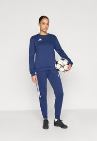 Σκούρα μπλε φούτερ και αντίστοιχα παντελόνια φόρμας με λευκές ρίγες Adidas. Το άτομο κρατάει μια ασπρόμαυρη ποδοσφαιρική μπάλα, φοράει μαύρα αθλητικά παπούτσια.