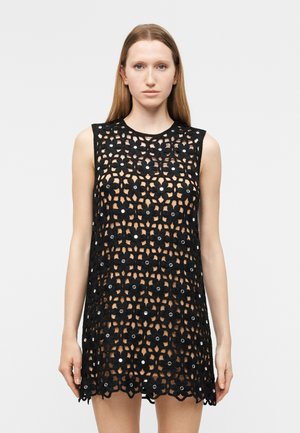 Γυναίκα που φοράει μαύρο πλεκτό φόρεμα χωρίς μανίκια με σχέδια λουλουδιών και μικρές κυκλικές λεπτομέρειες, στέκεται μπροστά σε απλό φόντο.