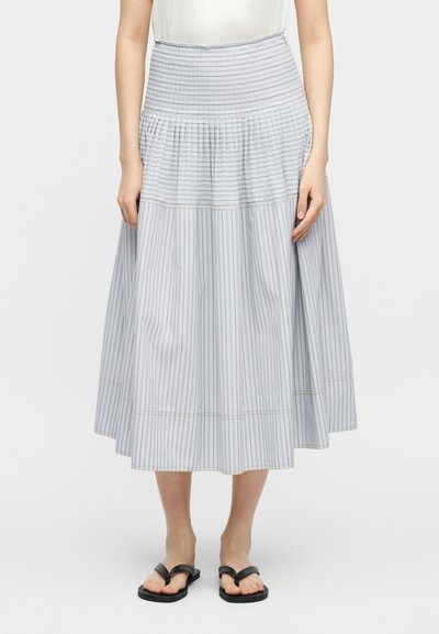 Femme portant une jupe midi blanche à rayures verticales fines, avec une large ceinture froncée, associée à des sandales plates noires, sur un fond uni.