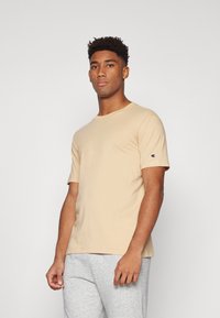 T-shirt en coton beige à manches courtes et col rond. Logo Champion en accent sur la manche gauche. Modèle portant un pantalon de jogging gris.