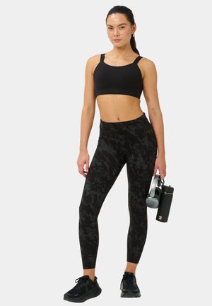 Vrouw staand in een zwarte sportbeha en een legging met print, met koptelefoon en een zwarte waterfles tegen een effen achtergrond.