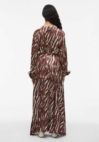 Femme se tenant de dos, portant une longue robe à motifs animaliers marron et beige avec des manches longues et des talons beiges.