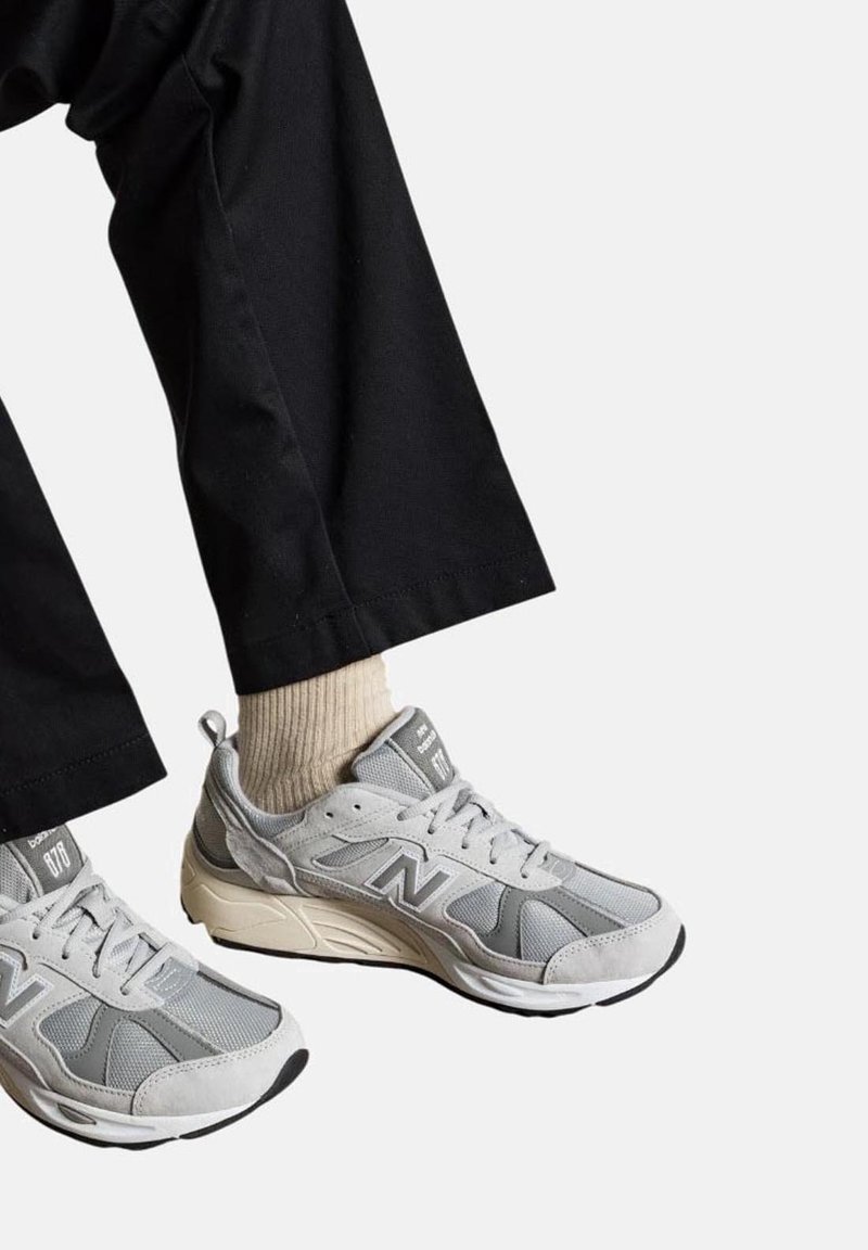 Graue Sportschuhe aus Mesh und Wildleder, mit einem auffälligen weißen "N"-Logo, kombiniert mit schwarzen Hosen und einer hellen Socke.
