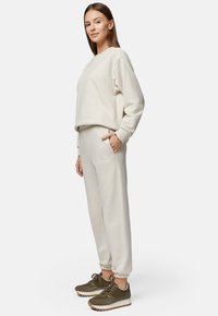 Beżowe spodnie dresowe z elastycznymi mankietami i kieszeniami, w połączeniu z dopasowaną bluzą z okrągłym dekoltem; model ma na sobie oliwkowe trampki.