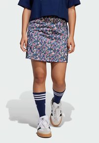 Φλοράλ φούστα σε σκούρο μπλε χρώμα με ίσια γραμμή, σε συνδυασμό με μπλε-λευκές ριγέ κάλτσες και λευκά αθλητικά παπούτσια με καστανές σόλες και μαύρες λεπτομέρειες.