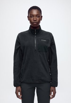 Modèle portant un pull polaire noir Columbia avec une fermeture éclair courte et une poche poitrine, devant un fond uni clair.