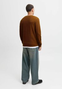 Brauner Waffelpullover mit langen Ärmeln, über einem weißen Hemd getragen; kombiniert mit lockeren, hellblauen Jeans und schwarzen Schuhen.
