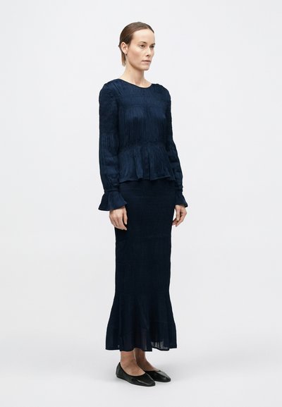 Femeie în picioare purtând o bluză plisată bleumarin cu manșete volanate și o fustă lungă asortată, încălțată cu balerini negri, pe un fundal alb simplu.