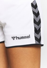 Shorts atléticos blancos con detalles en forma de chevron negros a lo largo de los lados y el logo "Hummel" en negro en la parte inferior izquierda. Textura suave.