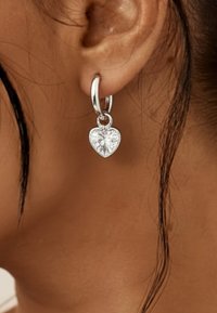 Orecchino a cerchio in argento con pendente a forma di cuore in cristallo trasparente, con superficie liscia e finitura lucida.