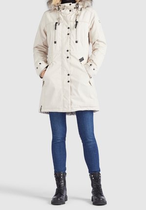 Kobieta w beżowej parkie sięgającej kolan z futrzanym kapturem, zapinanej na suwak i guziki, w połączeniu z niebieskimi dżinsami i czarnymi sznurowanymi botkami, stoi prosto.