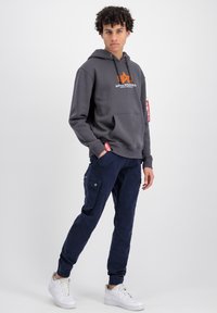 Felpa grigia con tasca frontale e logo, abbinata a pantaloni cargo blu navy con tasche. Sneakers bianche completano l'outfit.