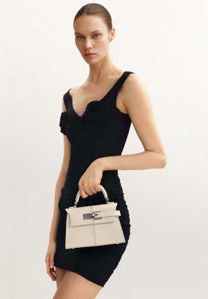 Femme en robe noire sans manches tenant un petit sac à main blanc sur un fond clair uni.