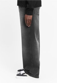 Black wide-leg jeans with a faded grey finish, featuring stitched detailing and a back pocket. Paired with black and white sneakers.
