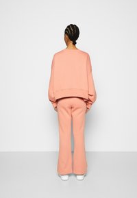 En person med flätat hår som bär en lös rosa sweatshirt och matchande byxor, står vänd mot en vit vägg och golv.