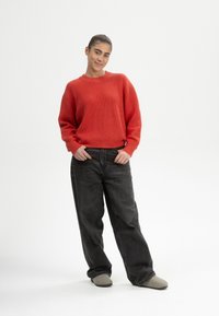 Rode geribbelde trui met vallende schouders, gedragen met loszittende donkergrijze jeans en lichtgrijze instappers. Op de achtergrond een effen witte achtergrond.