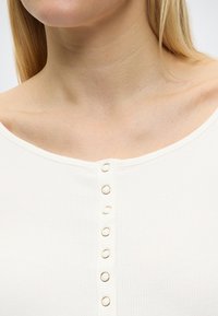 Κοντινό πλάνο στον λαιμό και το πάνω μέρος του στήθους ενός ατόμου που φοράει λευκό ριμπ μπλουζάκι με επτά μεταλλικά κουμπιά σε σχήμα δαχτυλιδιού κατά μήκος του κέντρου.